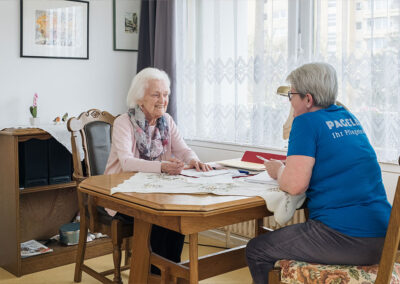 Pflegekraft und ältere Bewohnerin sitzen gemeinsam am Tisch in einer Wohngemeinschaft