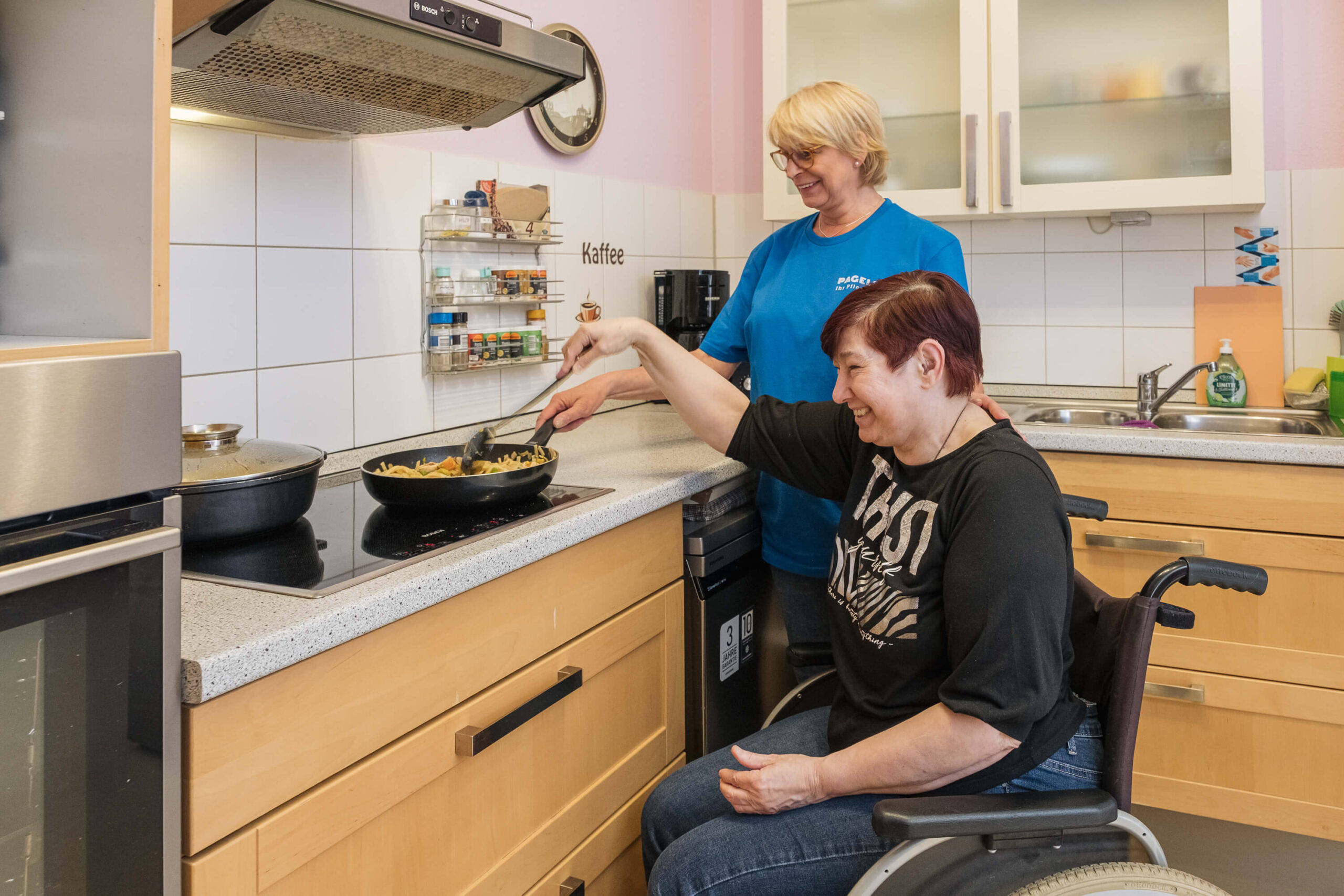 Eine Pflegekraft hilft einer Rollstuhlfahrerin beim Kochen in einer modernen Küche.