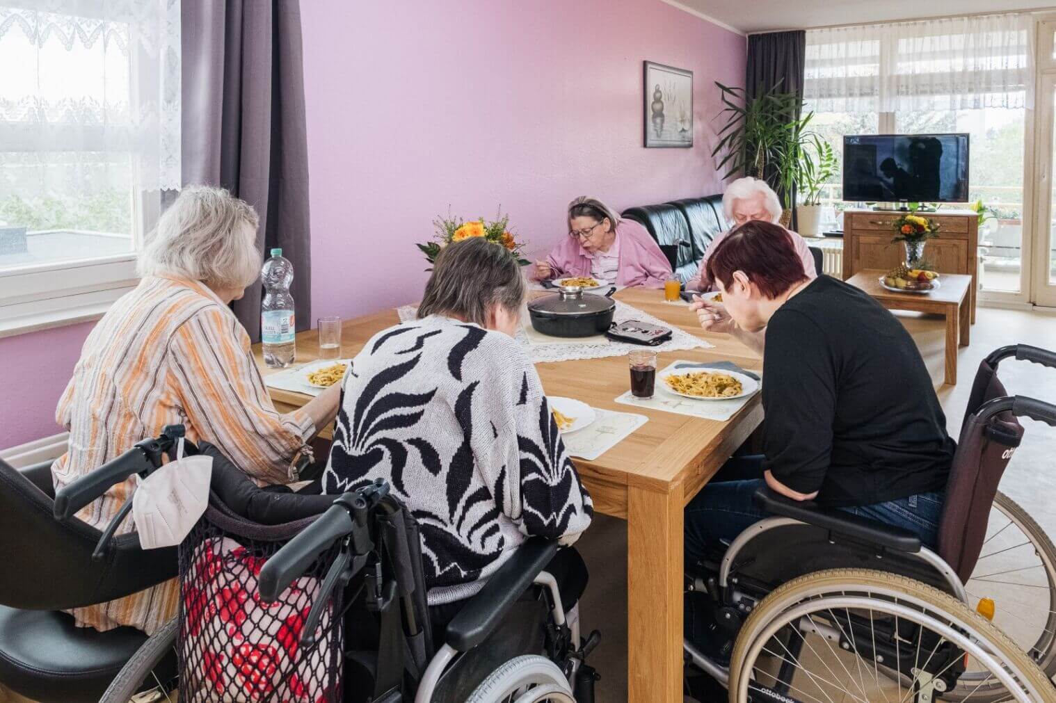 Ältere Menschen in einem hellen Esszimmer eines Seniorenheims sitzen gemeinsam am Tisch. Drei von ihnen sind in Rollstühlen und essen Pasta. Der Raum ist mit einer lila Wand und Pflanzen dekoriert, und es ist ein Fernseher im Hintergrund sichtbar.