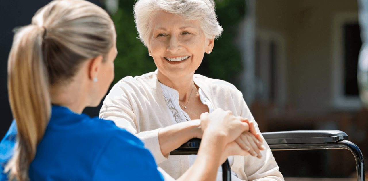 Pflegekraft sitzt mit einer Seniorin draußen und hält fürsorglich ihre Hand.