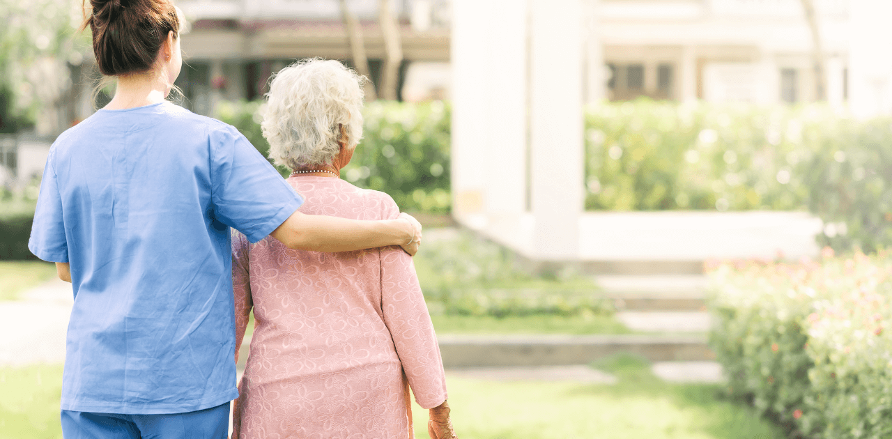 Pflegekraft begleitet eine Seniorin auf einem Spaziergang durch einen grünen Park.